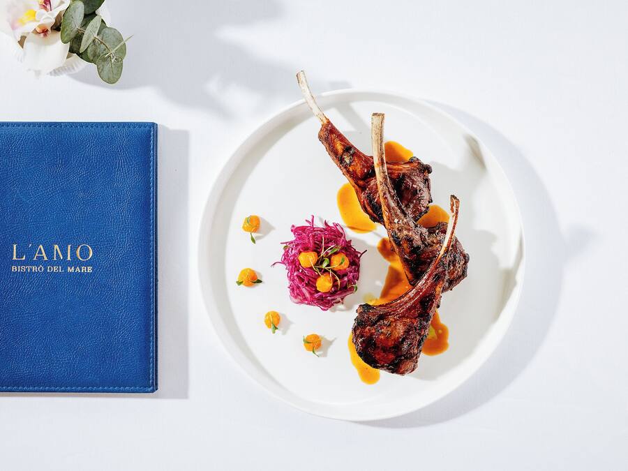 Un libro azul con el título "L'AMO BISTRO DEL MARE" junto a un plato blanco con chuletas de cordero y guarniciones.