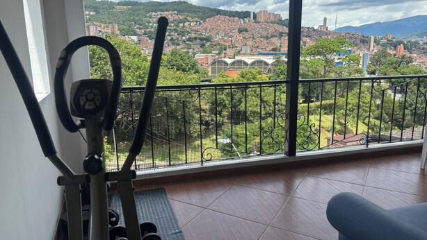 A gym with exercise equipment and a view of a cityscape through large windows. - Vista Hermosa Apartment In The Middle Of Medellin (Medellín)