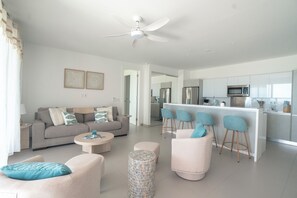 A modern living room with a sofa, chairs, a coffee table, and a patterned ottoman. A kitchen area with a bar and stools is visible in the background.