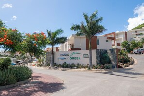 Señal de una comunidad residencial con la marca 'INDIGO GREEN', rodeada de palmeras y un camino pavimentado.