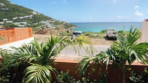 A coastal view with a parked van and a car, a building with a red and white facade, and a hillside with houses.