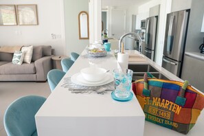 Una cocina moderna con electrodomésticos de acero inoxidable, una zona de comedor con mesa blanca, sillas azules y un bolso colorido con la etiqueta 'St. Maarten'.