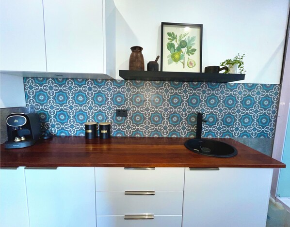 Una cocina con encimera de madera, una cafetera, dos tazas, un lavamanos y una planta decorativa en una estantería.