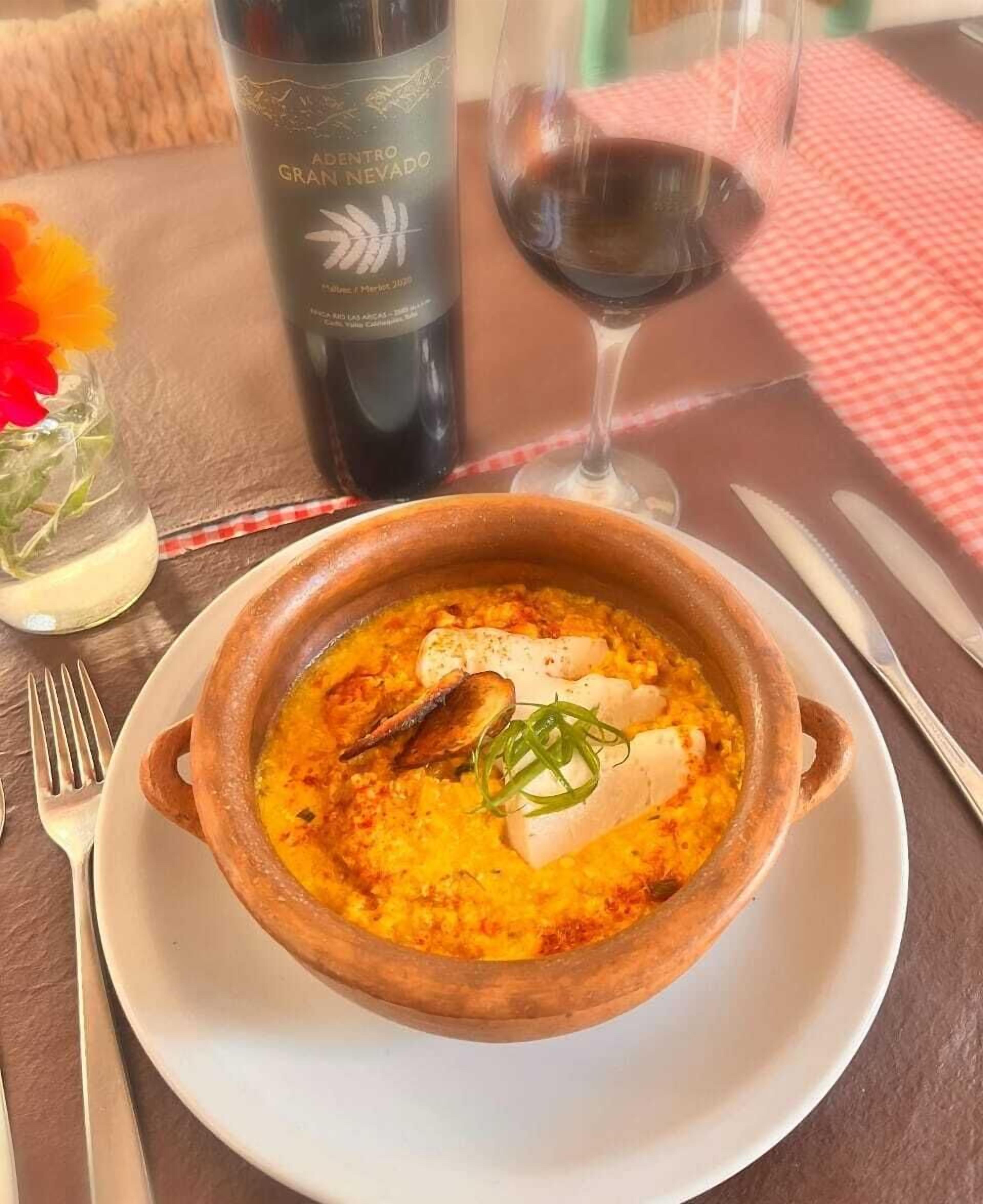 A bowl of seafood soup on a white plate, accompanied by a wine glass and a bottle of wine on a table.