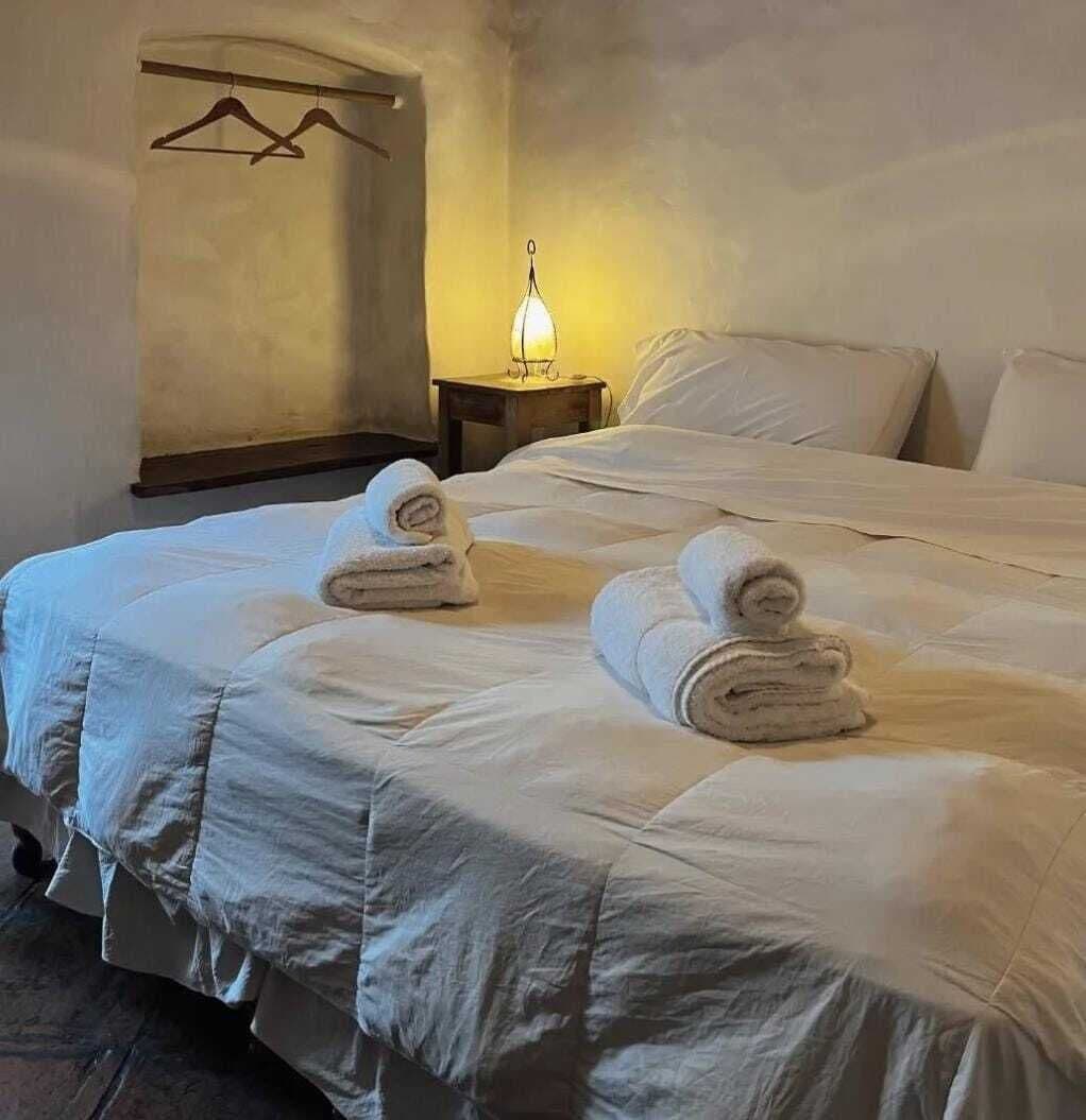 Habitación de hotel con una cama grande, dos toallas enrolladas sobre la cama, una lámpara de noche y un perchero con perchas.