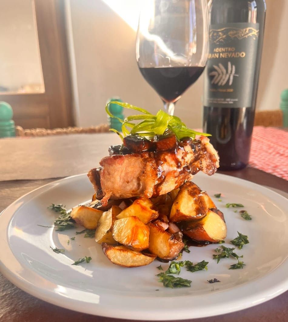 A plate of roasted meat and potatoes garnished with herbs, with a bottle of wine and a glass in the background.