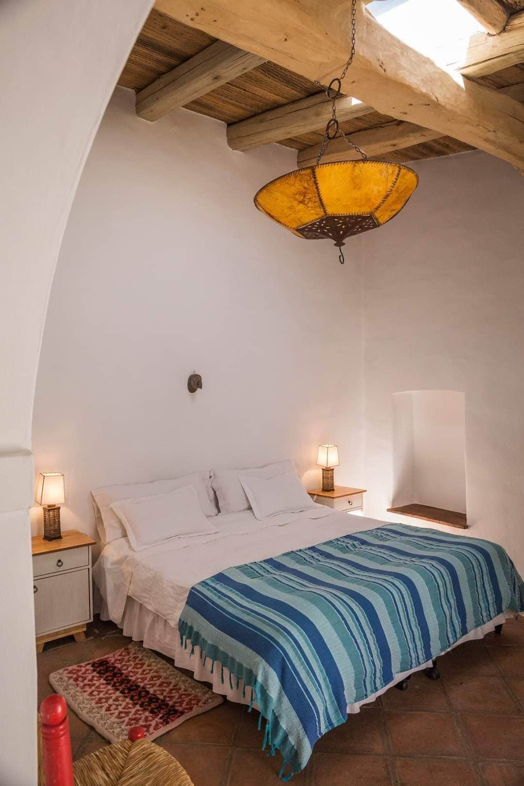 Un dormitorio con una cama, dos mesitas de noche con lámparas, un aplique colgante y una silla roja.