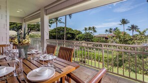 A dining area with a table set for four, overlooking a lush garden with palm trees and a view of a residential area. - Spacious Family Retreat! Ocean View Sunsets + Pool, Spa & Beach Club (Waimea)