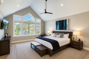 A bedroom with a large bed, two bedside tables, a ceiling fan, and a view of trees through large windows.
