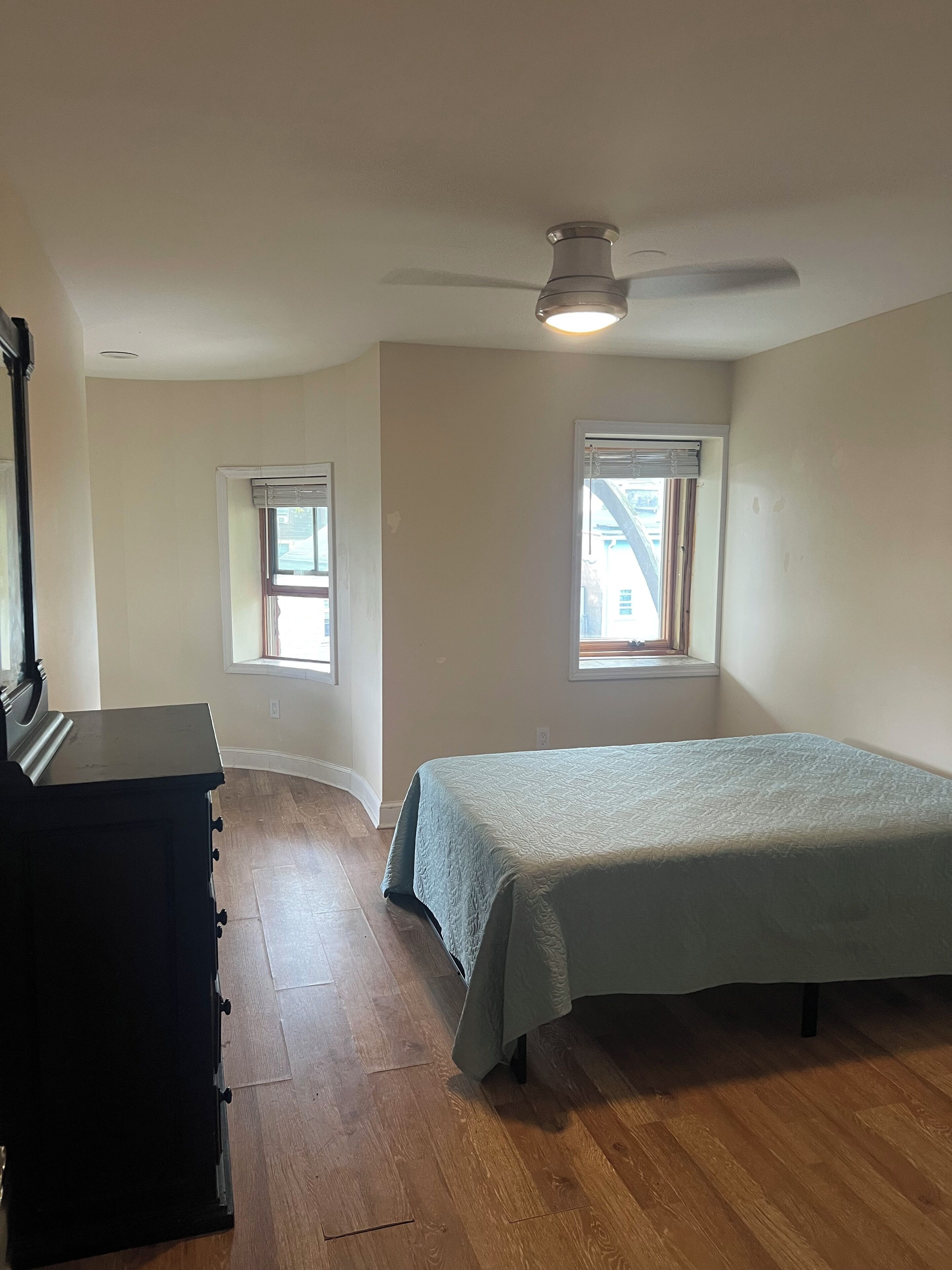 Une chambre à coucher comprenant un lit, une commode, deux fenêtres et un ventilateur de plafond.