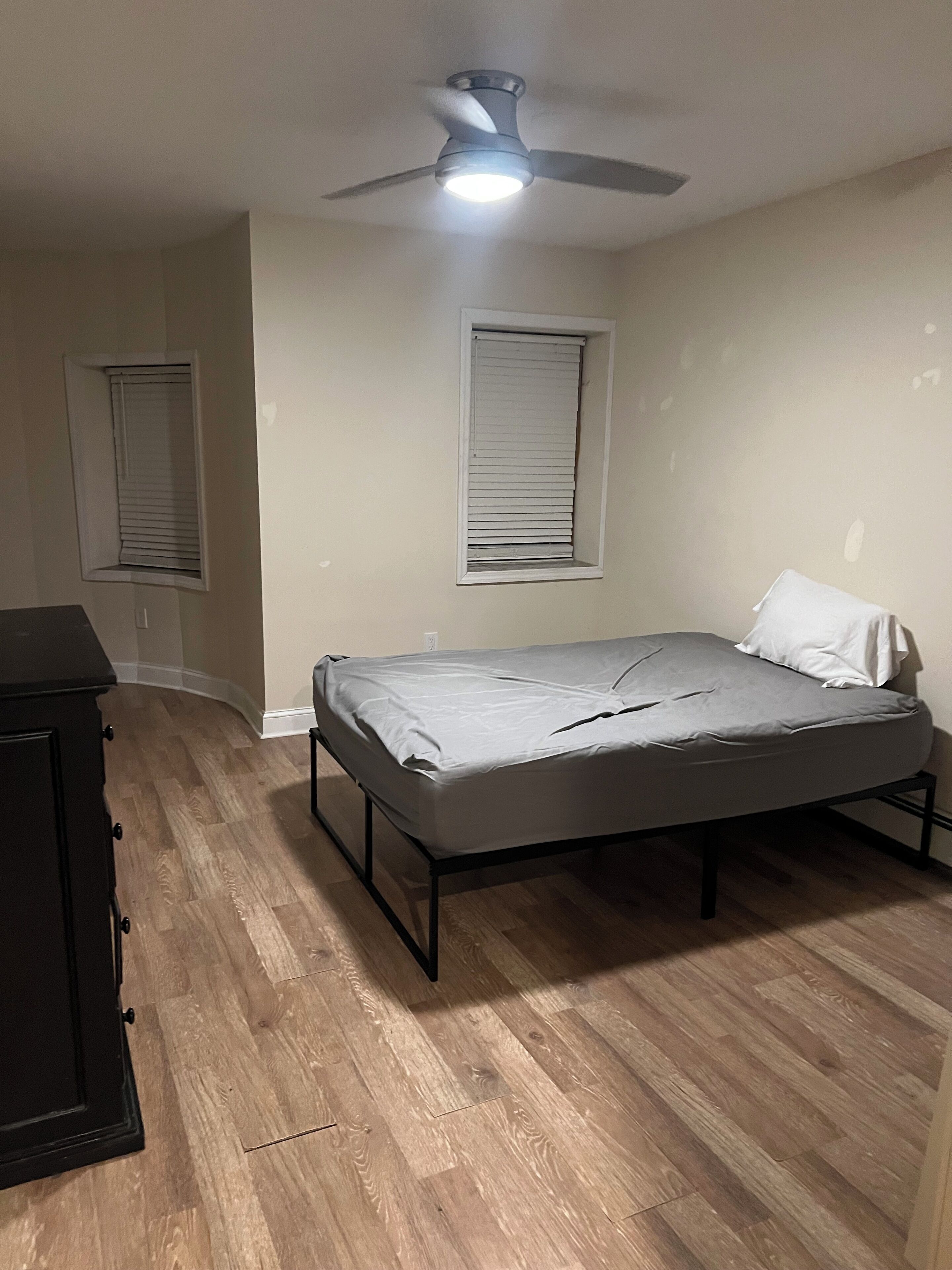 Een slaapkamer met een enkel bed, een plafondventilator en twee ramen met jaloezieën.