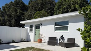 A single-story house with a white exterior, a green door, and a window with blinds. There is a potted plant and a bicycle parked outside.