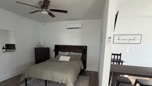 A bedroom with a bed, a ceiling fan, an air conditioning unit, a bedside table with a lamp, and a wall-mounted sign.