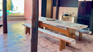 Ein überdachter Außenbereich mit Holztisch und Bänken, einem Fliesenboden und einem Kühlschrank.