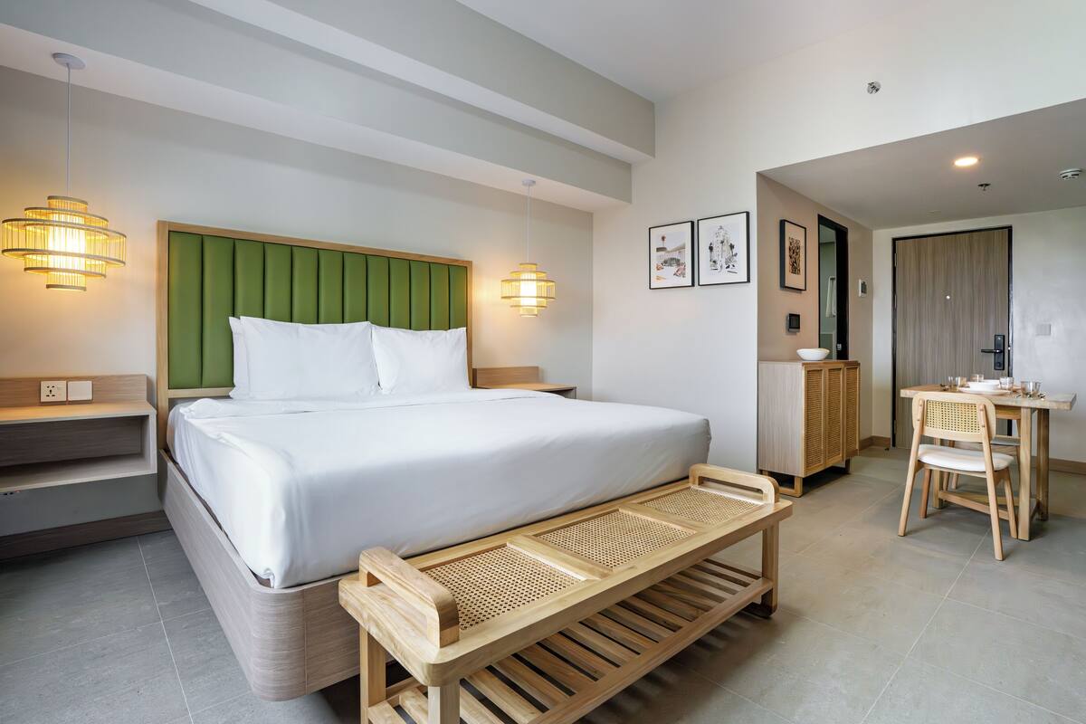A modern hotel room with a large bed, bedside tables, a wooden bench, a dining table with chairs, and wall-mounted lights.