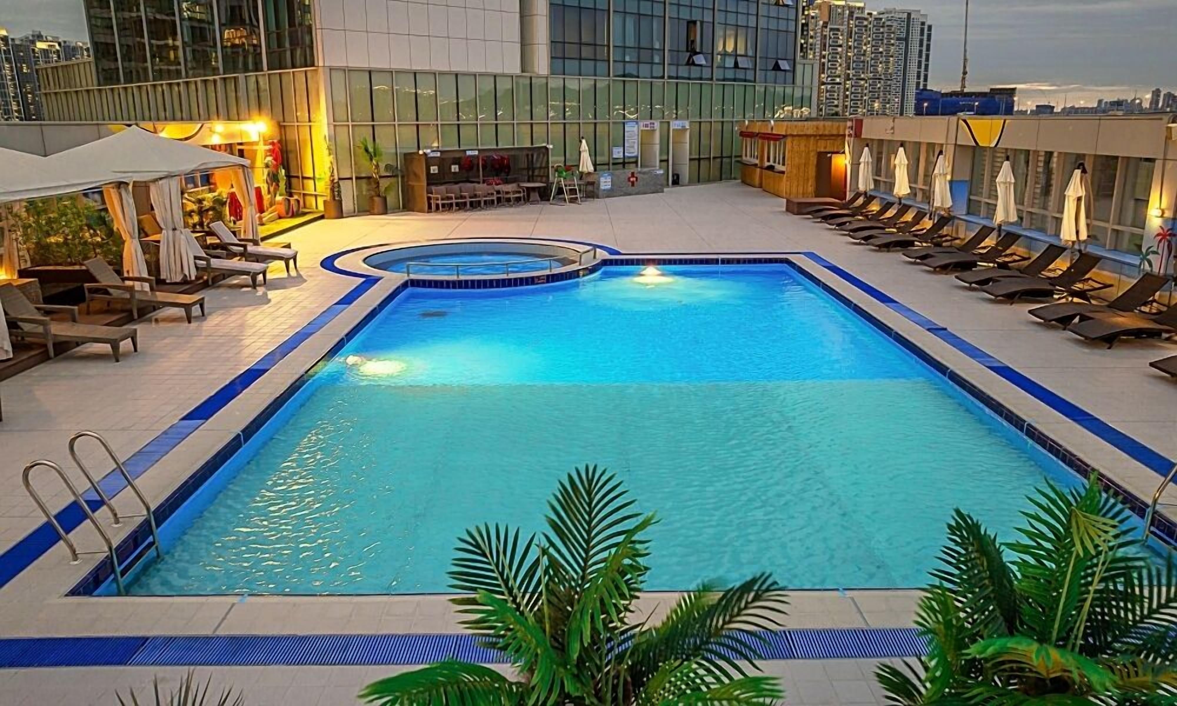 A rectangular pool with a blue border, surrounded by lounge chairs and umbrellas, with a cityscape in the background.