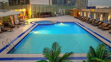 A rectangular pool with a blue border, surrounded by lounge chairs and umbrellas, with a cityscape in the background.