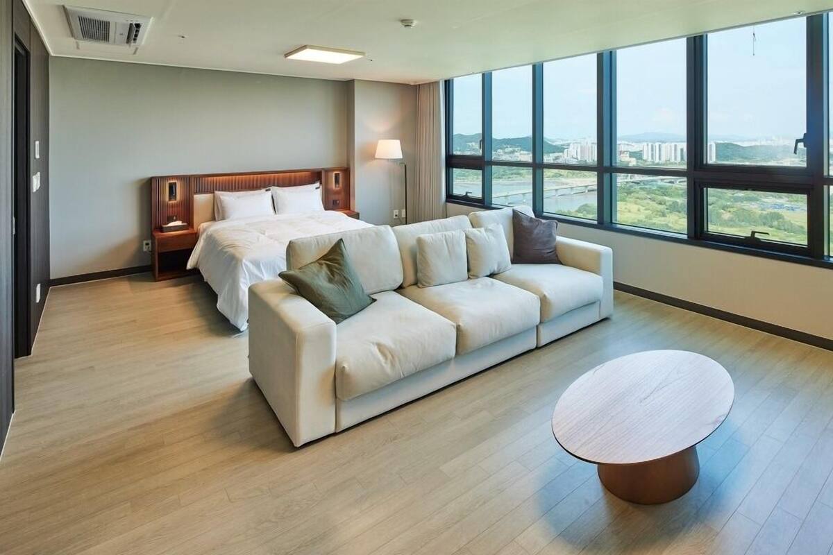A hotel room with a bed, a sofa, a coffee table, and large windows offering a city view.