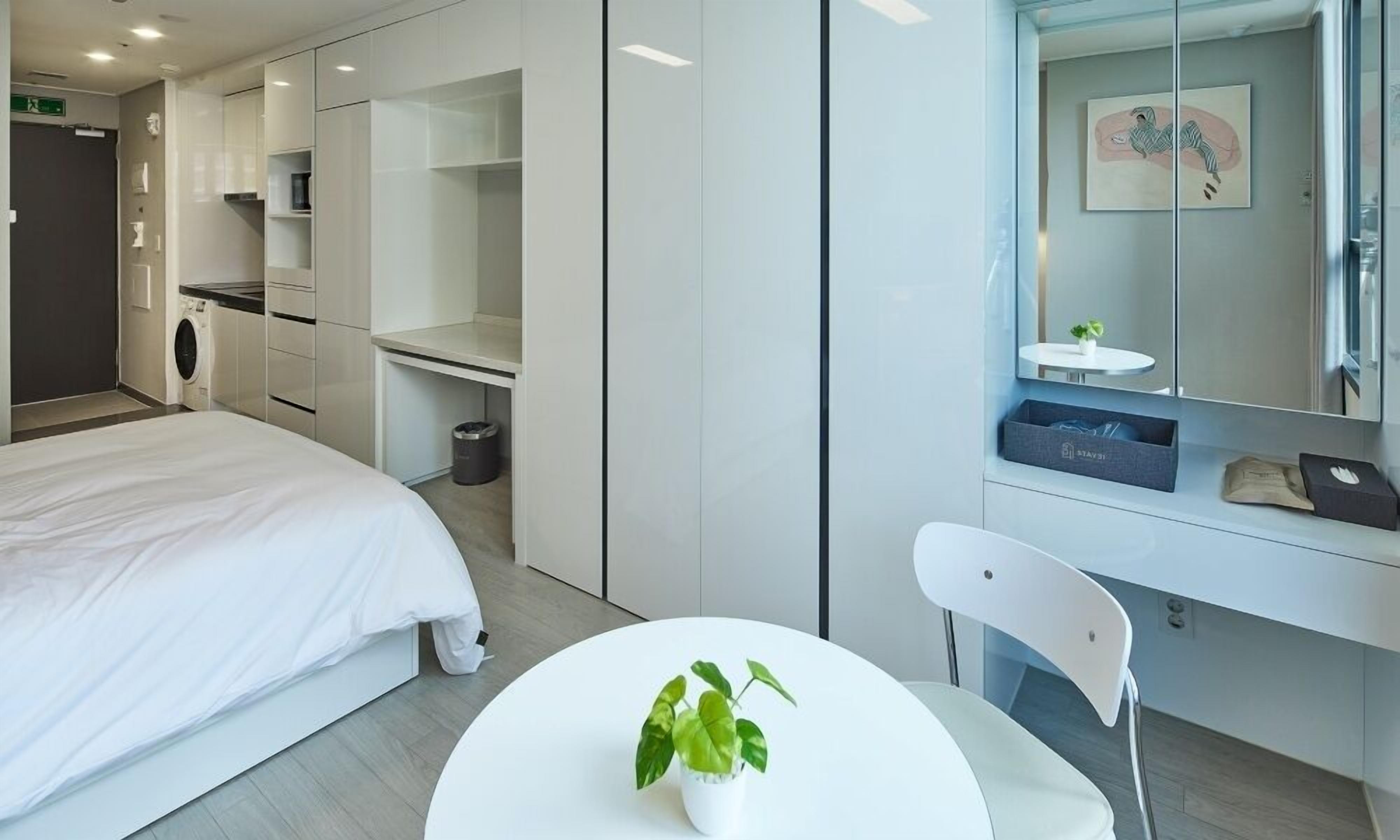 A modern hotel room with a bed, a small table with a plant, a desk with a computer, and a bathroom with a mirror and sink.