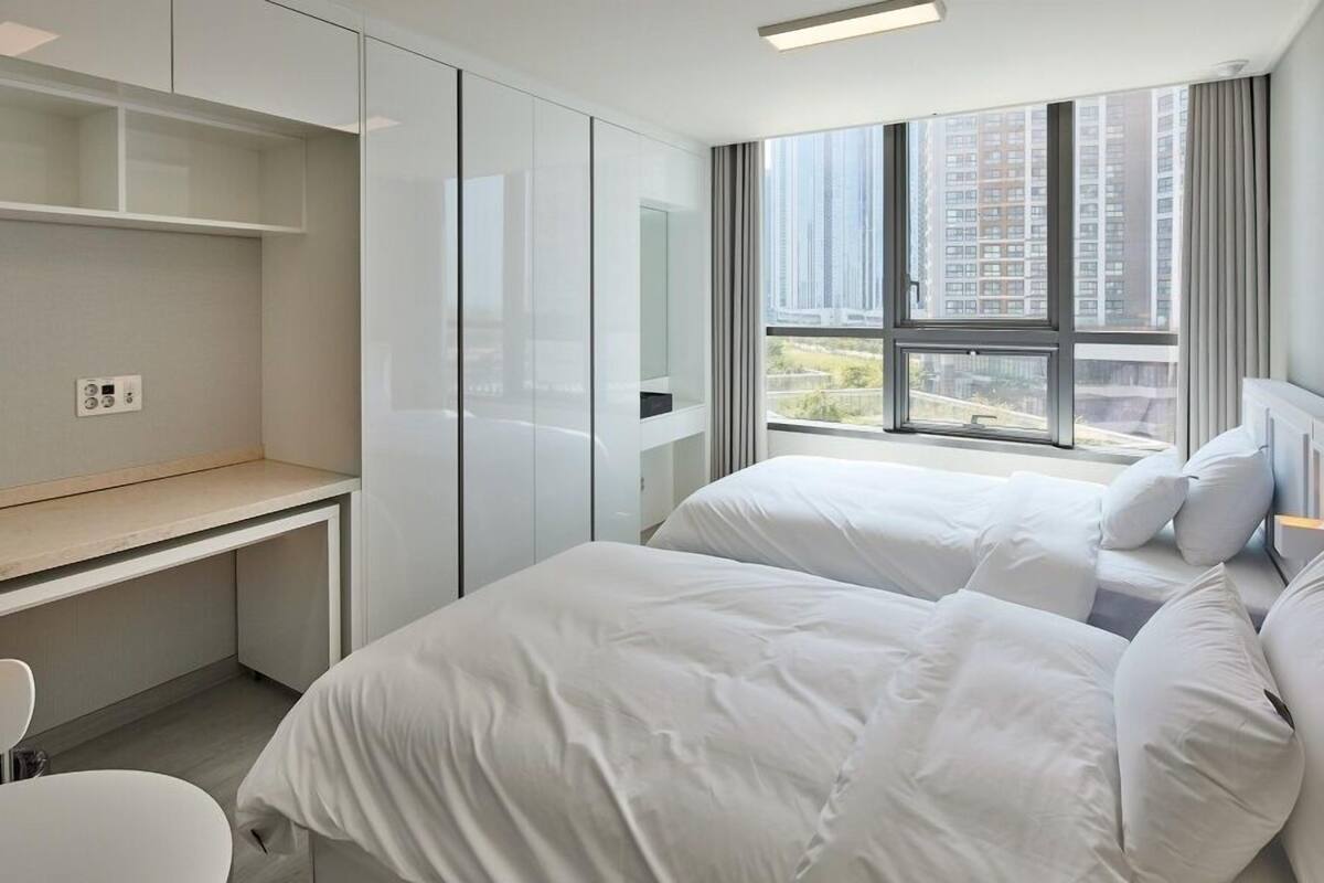 A modern hotel room with a large window, a desk, and two beds.