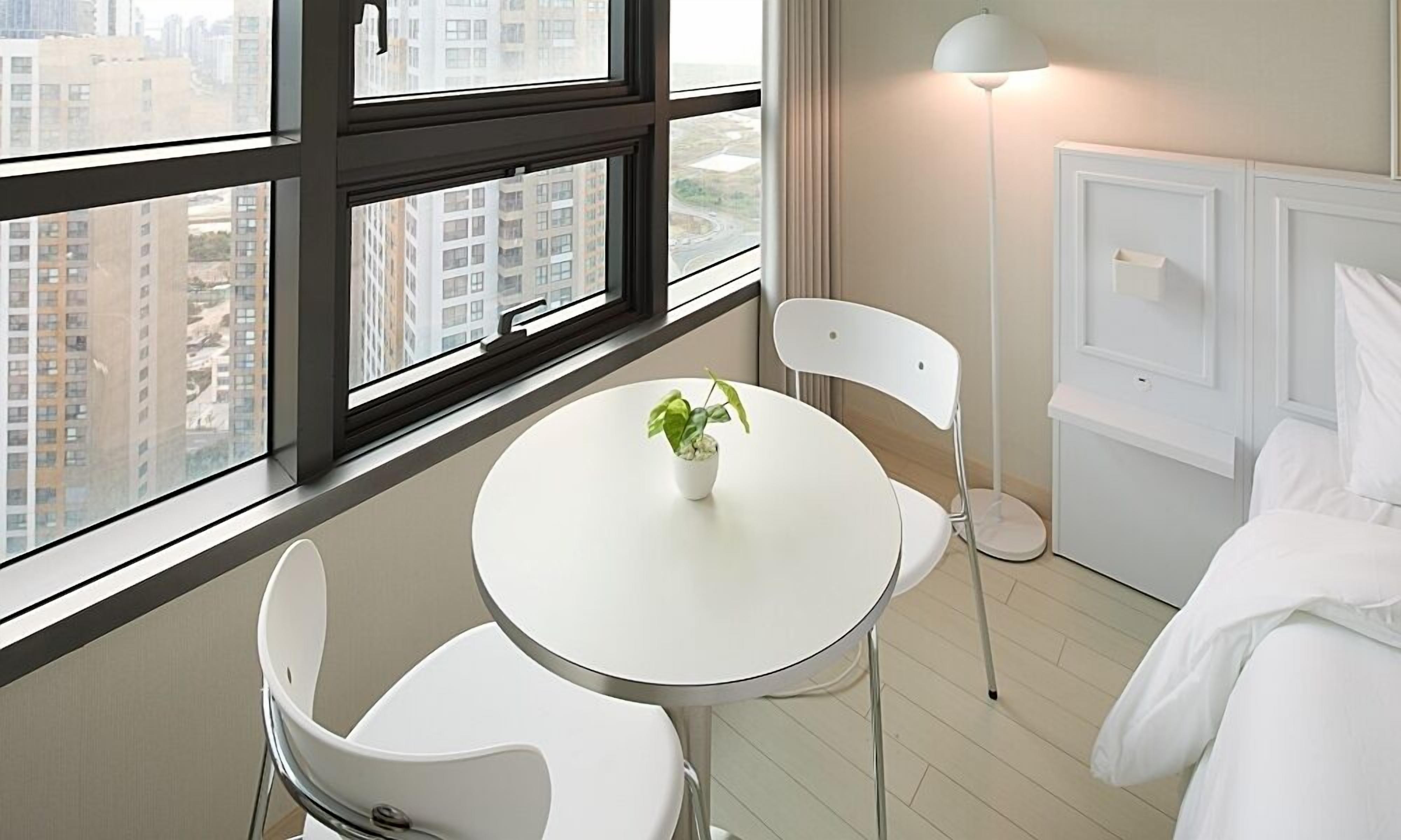 Une chambre d’hôtel avec une table ronde blanche, une plante en pot, une chaise blanche, un lit avec une literie blanche et une vue sur les bâtiments à travers de grandes fenêtres.