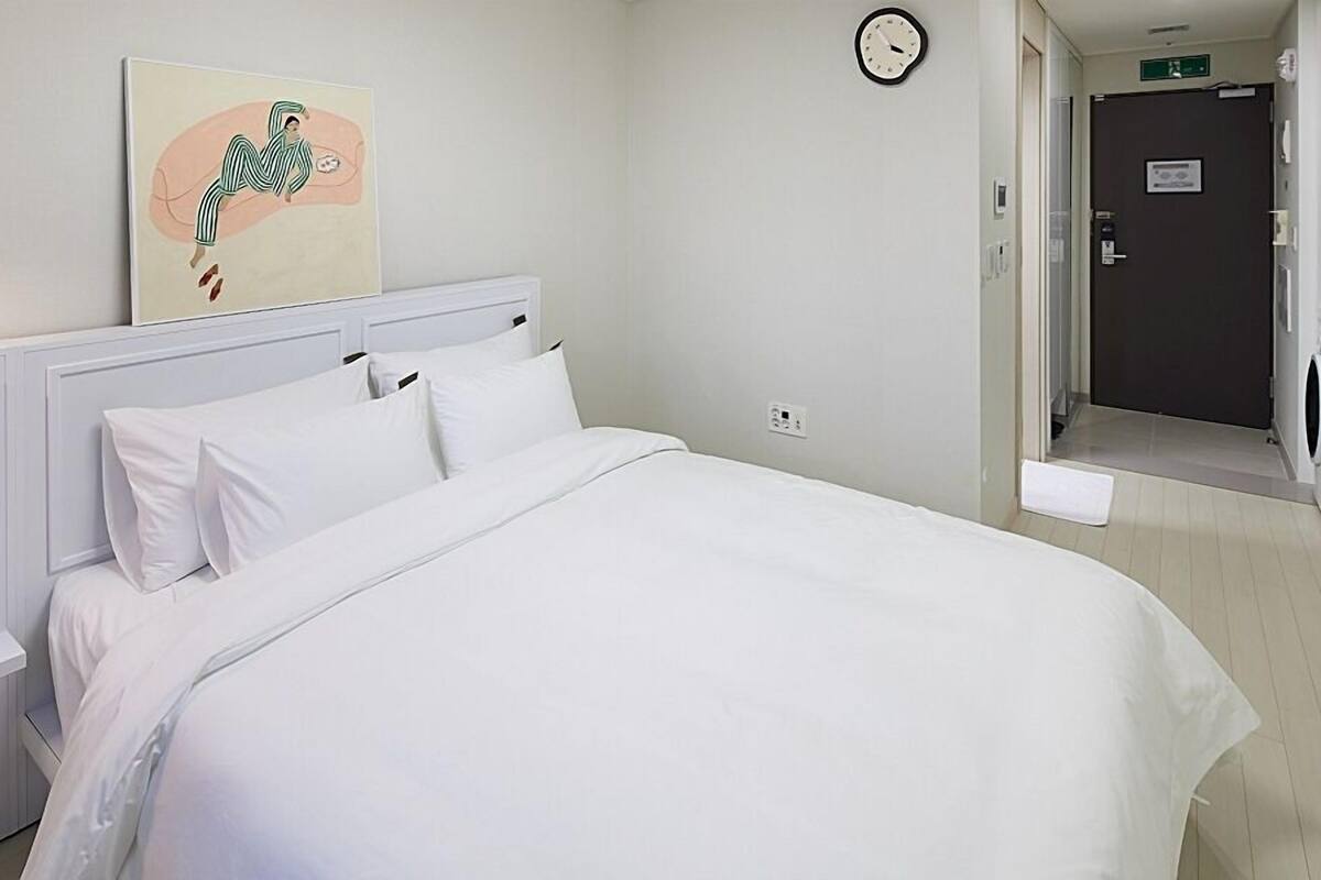 A hotel room with a bed, white bedding, a painting on the wall, a clock, and a door leading to a laundry area.