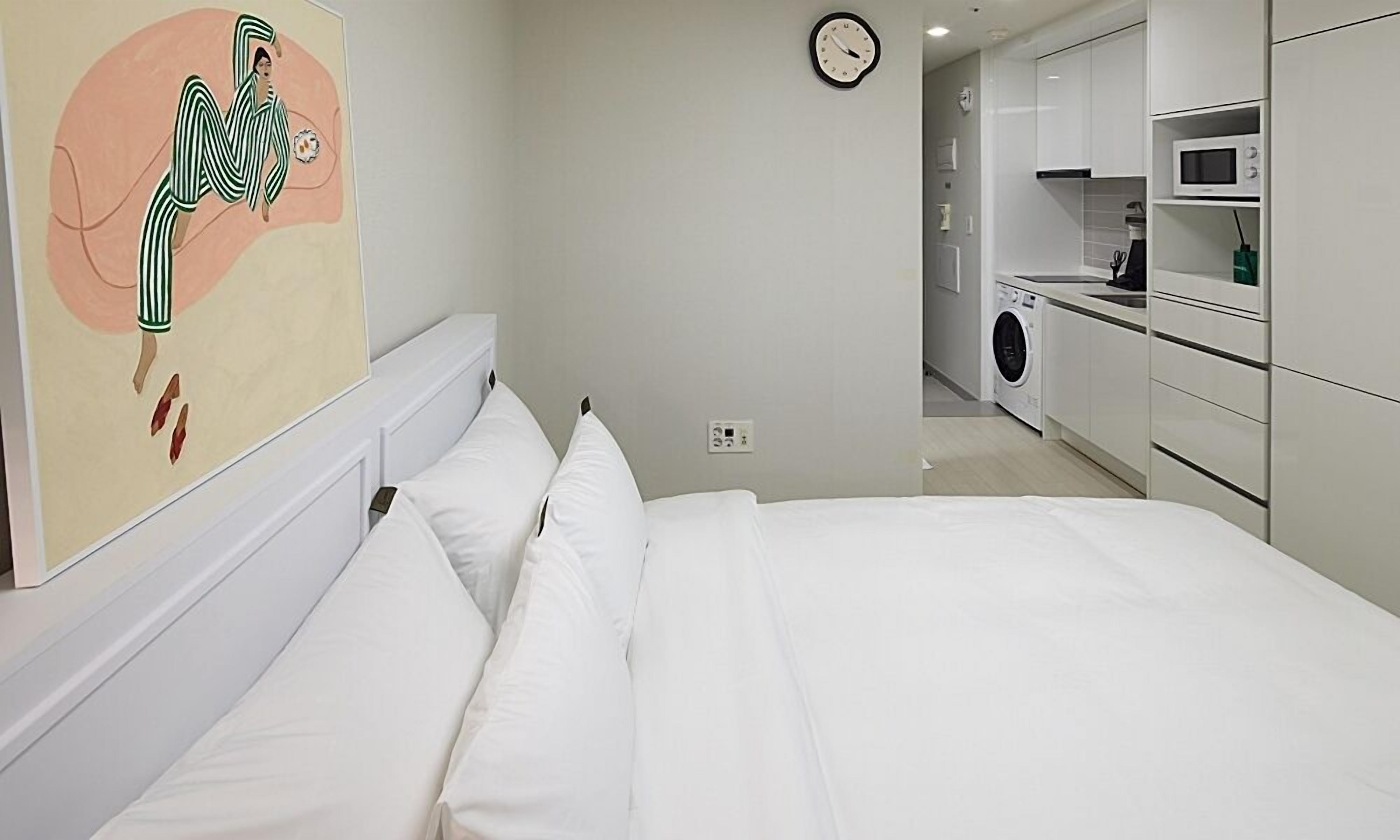 A bedroom with a bed, a wall clock, a painting, and a laundry area with a washing machine.
