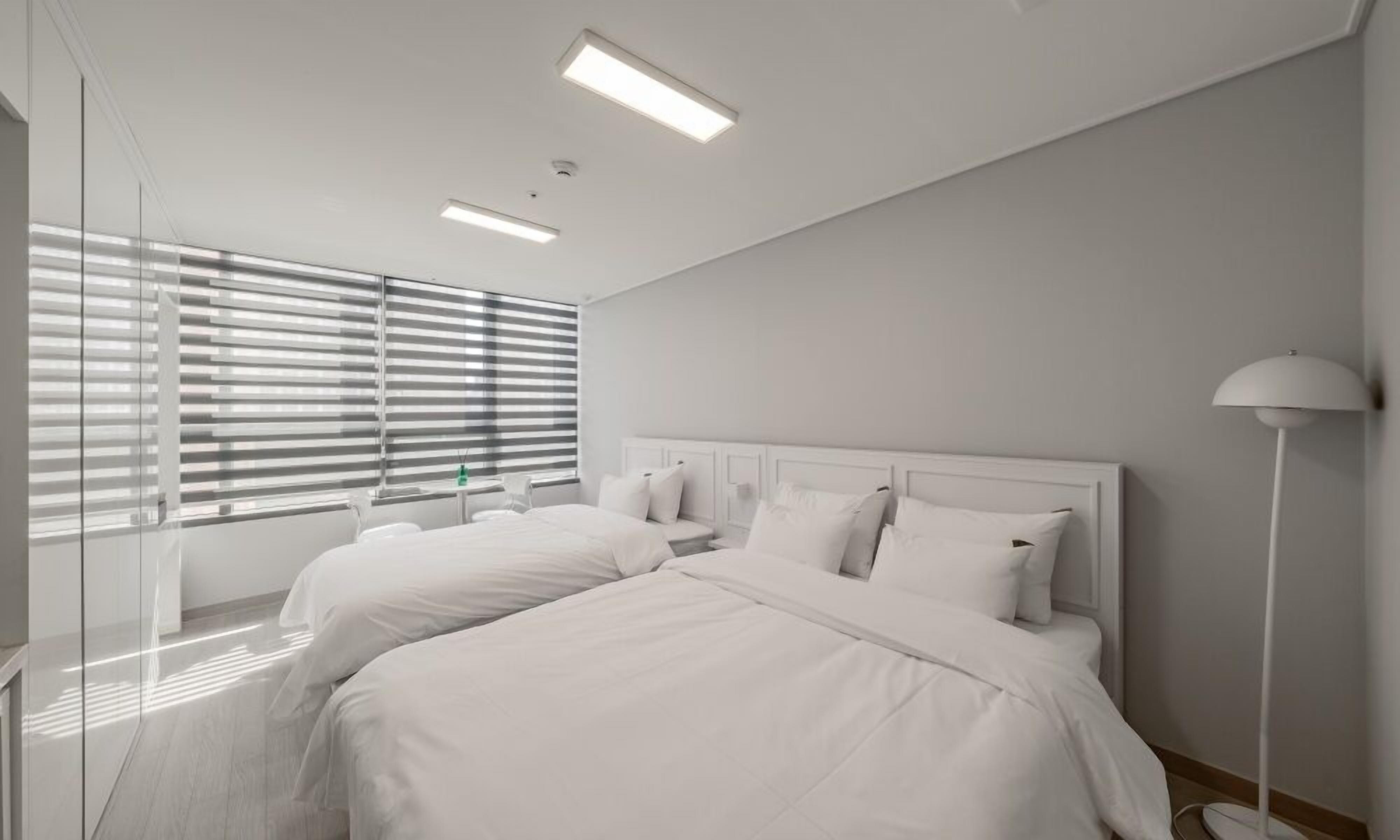 A modern hotel room with two beds, a large window with blinds, a bedside lamp, and a mirror.