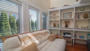 Una sala de estar acogedora con un sofá, una estantería y vistas a árboles a través de las ventanas.