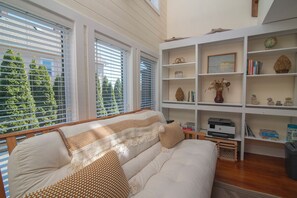 A cozy living room with a sofa, a bookshelf, and a view of trees through the windows.