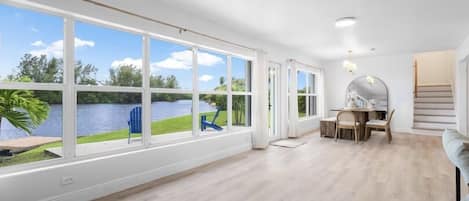 A spacious room with large windows, a wooden floor, a dining area with a table and chairs, and a view of a lake.