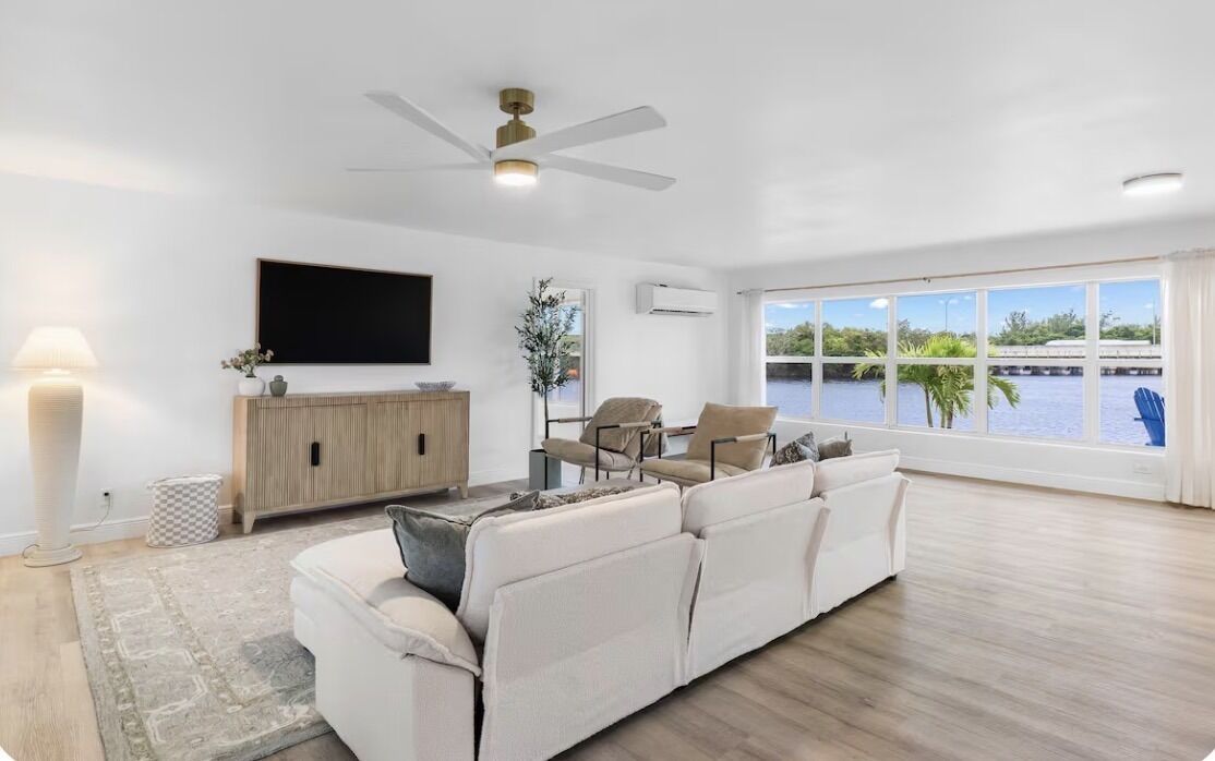 A spacious living room with a white sofa, a TV, a ceiling fan, and a large window with a view of a body of water.
