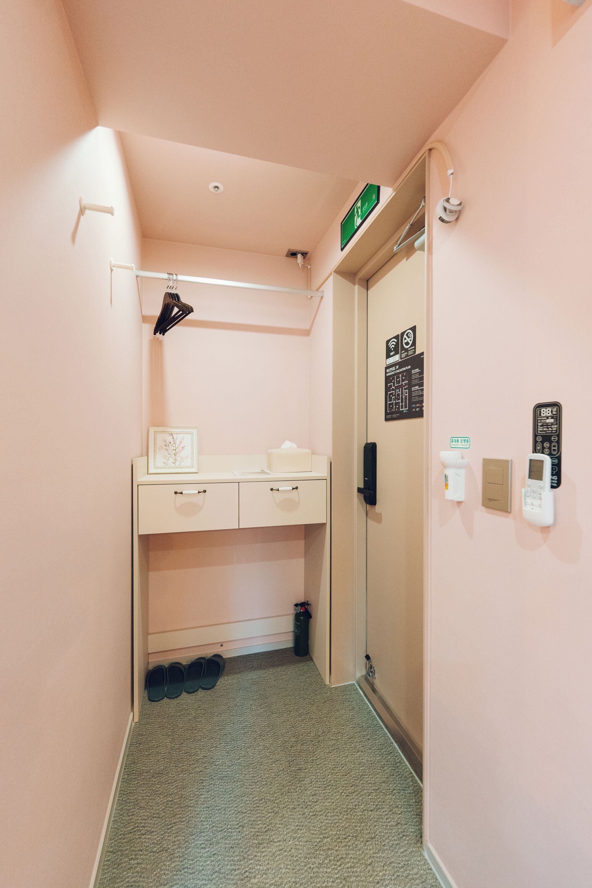 Um corredor com carpete bege, uma escrivaninha branca com gavetas, um quadro emoldurado, uma luminária suspensa, uma porta com identificação verde e um telefone fixado na parede.