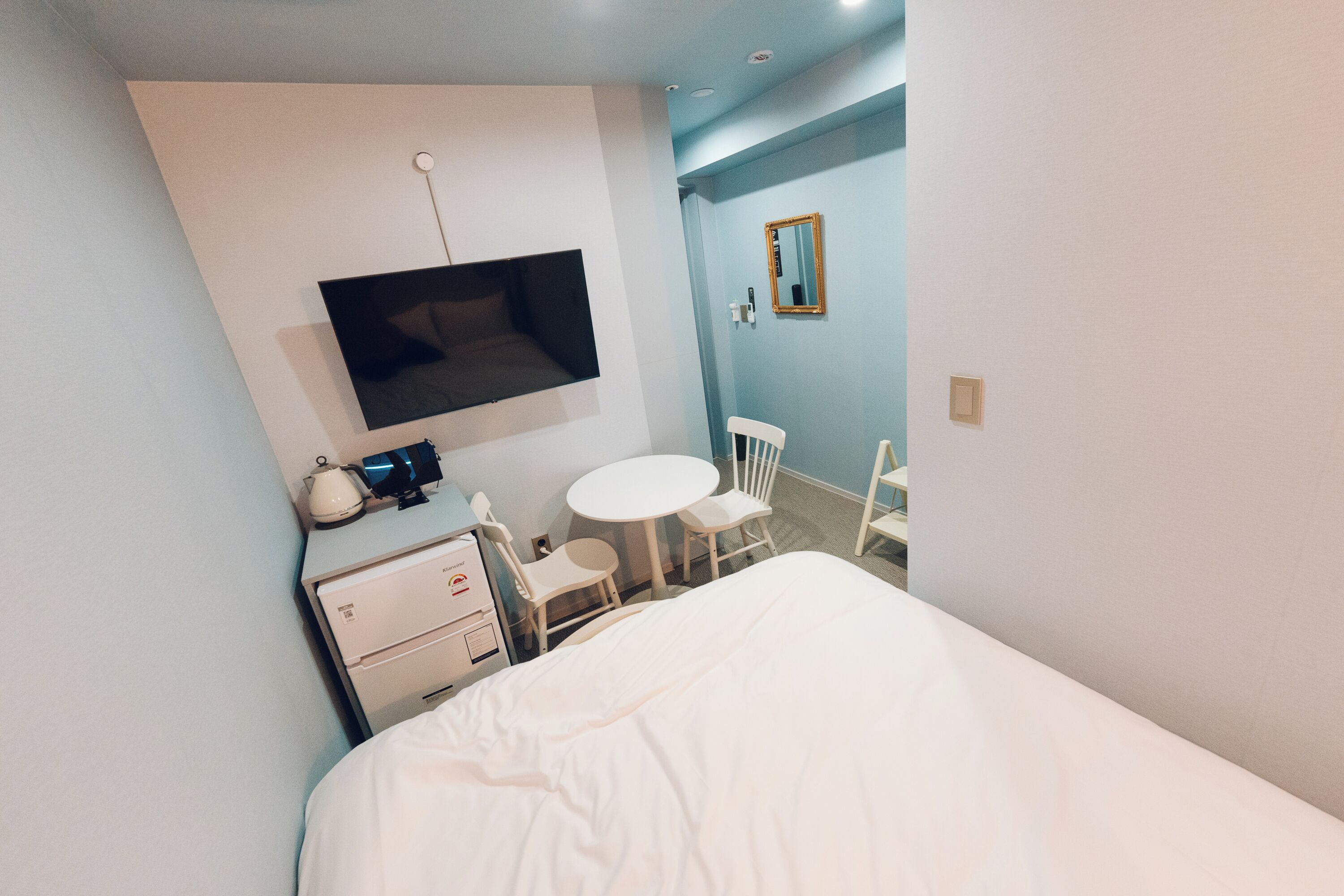 A hotel room with a bed, a desk, a chair, a small table, a TV mounted on the wall, and a mirror.