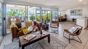 Un'ampia zona giorno con un divano in pelle marrone, un tavolino da caffè e un tavolo da pranzo per quattro persone. Grandi vetrate conducono a un balcone con vista sul mare.