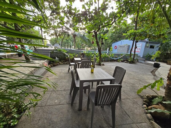 An outdoor patio area with a table and chairs, surrounded by greenery and a blue building in the background. - Kawan Beach House and Sanctuary (Lian)