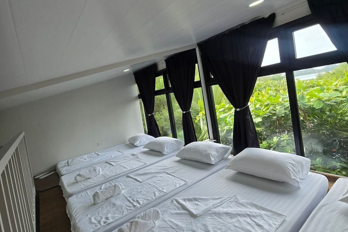 Ein Hotelzimmer mit zwei Betten, großen Fenstern mit schwarzen Vorhängen und Blick auf Grünfläche.