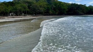 Ein Strand mit klarem Wasser, einem sandigen Ufer und einer Hügelkette mit dichter Vegetation im Hintergrund.