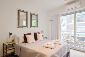 Habitación de hotel con cama, dos mesitas de noche, una lámpara de pie, una planta en maceta y vistas al balcón exterior.