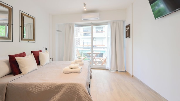 A hotel room with a large bed, a flat-screen TV, a balcony with outdoor furniture, and a view of a building.