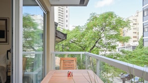 A balcony with a wooden table and chair, overlooking a cityscape with buildings and trees.