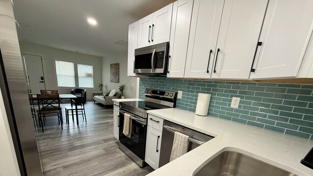 A modern kitchen with white cabinets, a stainless steel sink, a microwave, and an oven. There is a dining area with a table and chairs in the background.