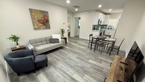 A modern living room with a grey sofa, a dining area with a wooden table, and a kitchen in the background.