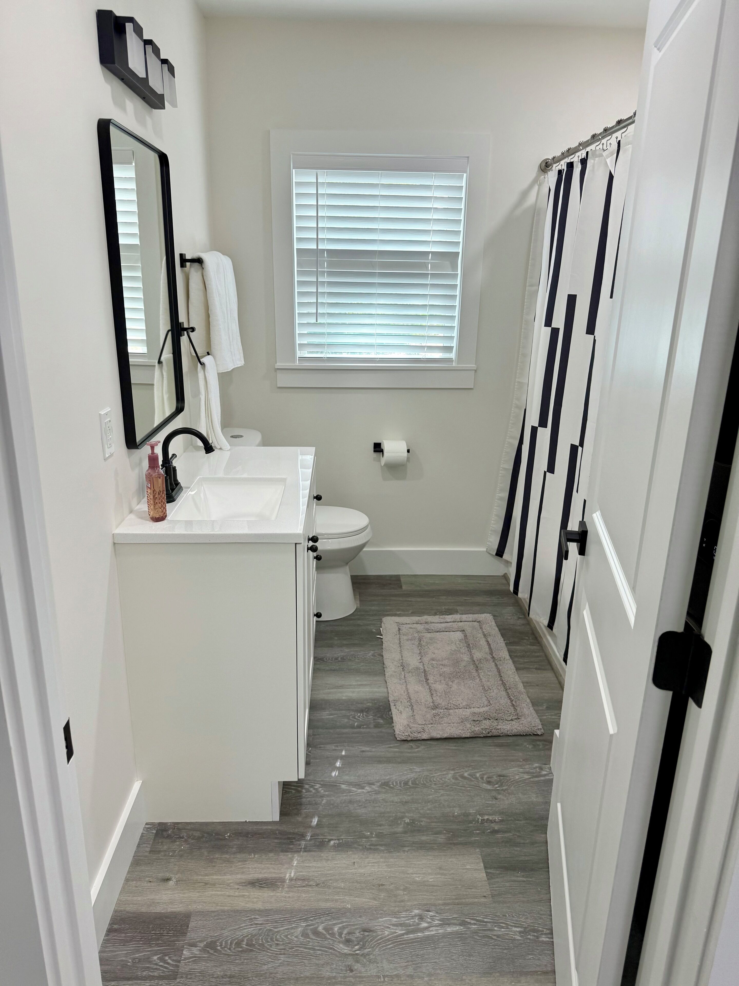 A bathroom with a white sink, mirror, and toilet, a window with blinds, and a gray rug on the floor.
