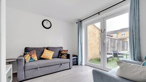 Una sala de estar moderna con un sofá gris, cojines estampados, un reloj de pared y un ventanal corredero que da a un jardín.