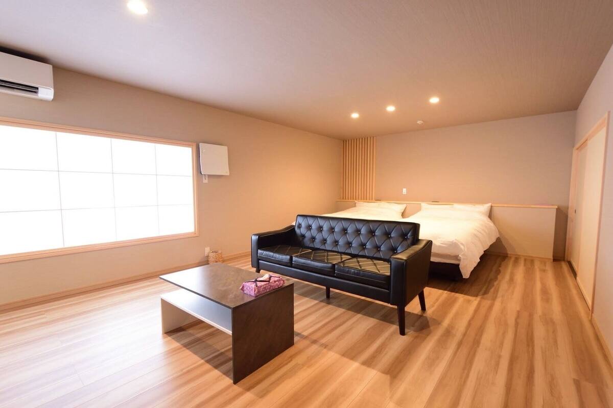Ein Schlafzimmer mit einem Bett, einem schwarzen Sofa, Holzboden, einem Fenster und einer Klimaanlage.