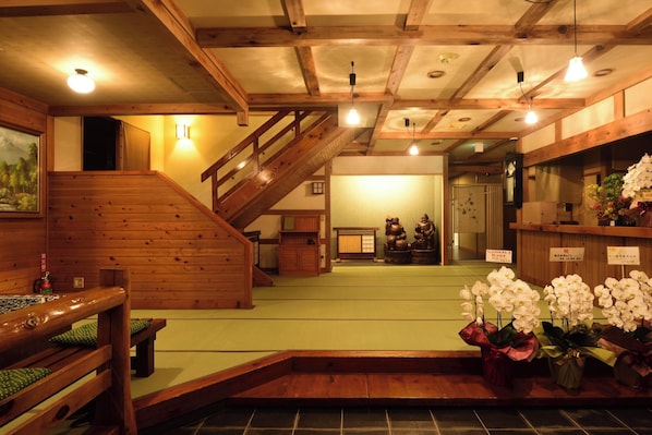 Un intérieur japonais traditionnel avec un sol recouvert de tatamis, des poutres en bois, un escalier et un espace d’accueil agrémenté de compositions florales.