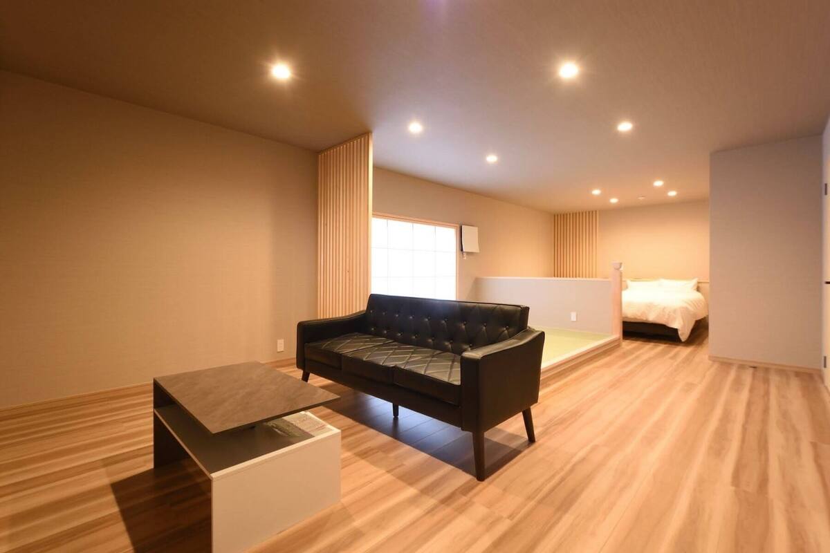 Ein modernes Wohnzimmer mit einem schwarzen Sofa, einem Holztisch und einem Bett im Hintergrund.