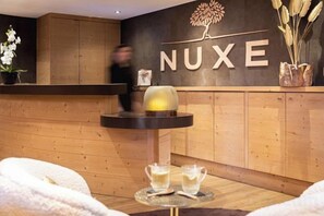 Un espace d’accueil avec un comptoir en bois, une enseigne fixée au mur portant l’inscription « NUXE », et une petite table avec deux verres et une théière.