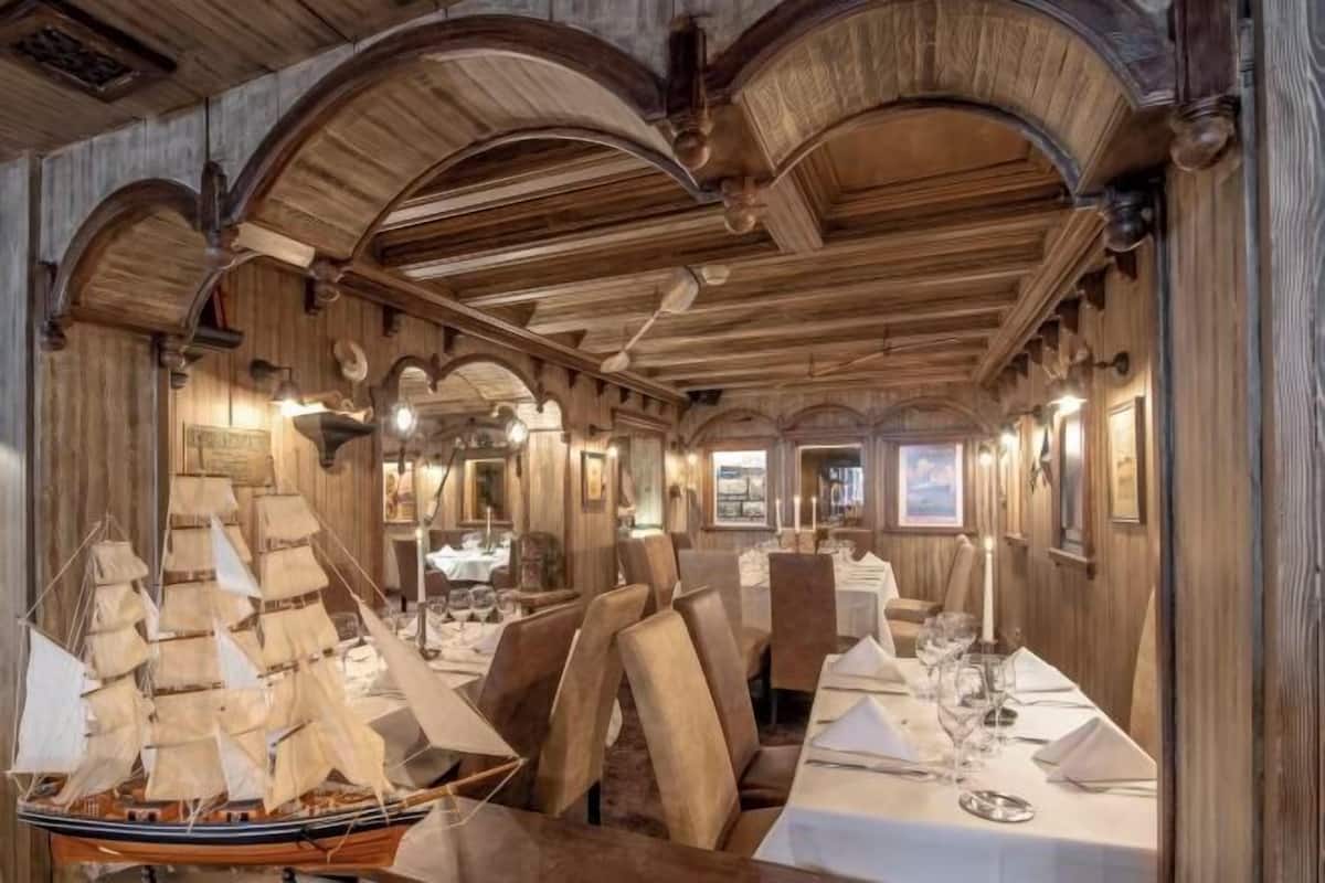 Un comedor con paneles de madera, techos abovedados y un modelo de barco. Las mesas están dispuestas con manteles blancos y velas.
